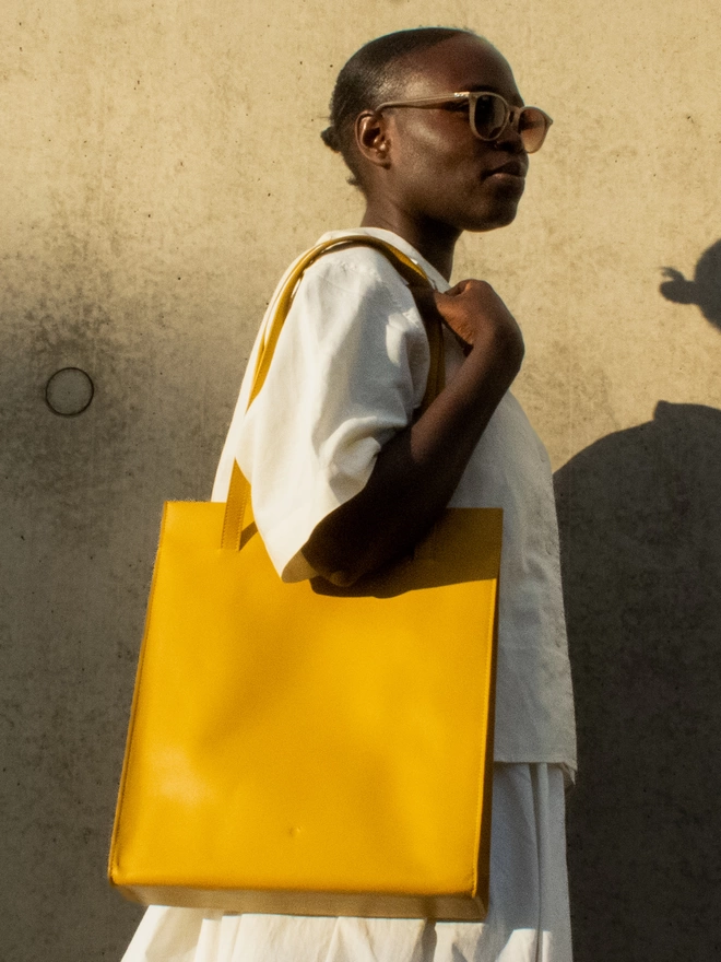 Amber Yellow Tote Bag worn on a oversized white shirt and skirt. The model is of a beautiful darker complexion with amber sunglasses against a serpia toned wall