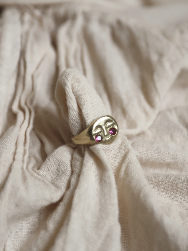 Image of a gold moon face ring with ruby cheeks on a piece of cream linen type fabric 