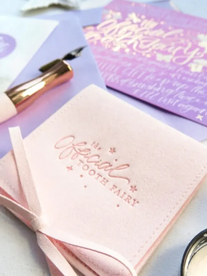 Close-up of a blush pink suede pouch with the "Official Tooth Fairy" logo embossed in pink. A rose gold calligraphy pen and a pink-lilac notecard sit behind it.