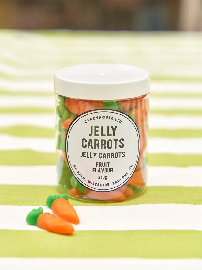 A clear jar with a white lid full of jelly carrot sweets placed on a green and white stripped tablecloth, there are a couple of jelly carrots next to the jar 
