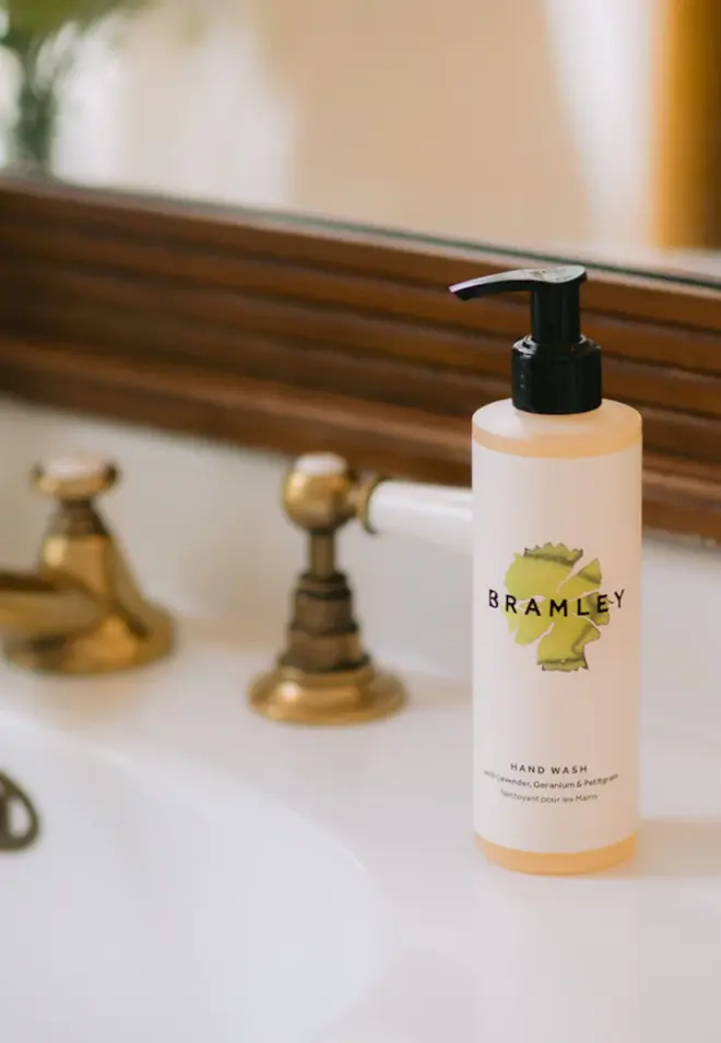 bottle of pale orange liquid, with white label and green logo, and black pump-lid. sitting on a white sink, with gold faucet and taps, and a brown-framed mirror in background