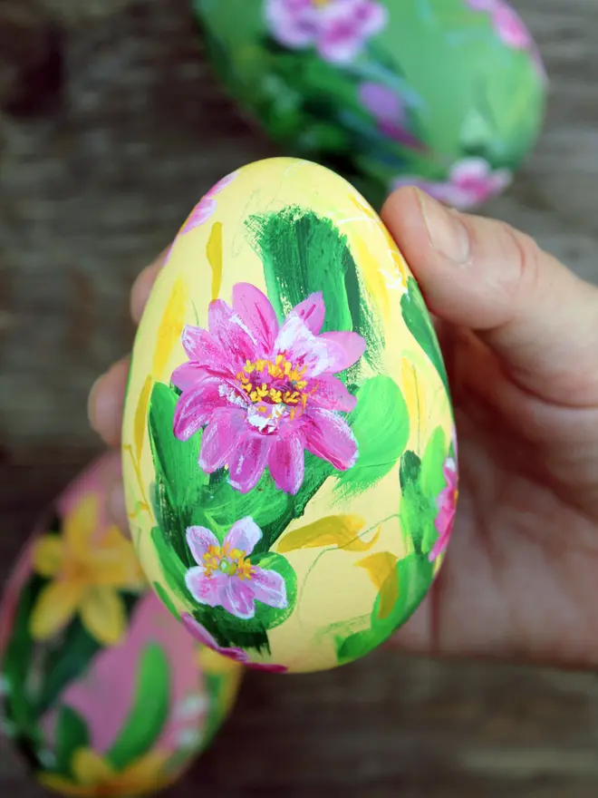 Hand painted floral wooden egg pale buttery yellow background adorned in pink zinnias and pink japanese anemone flowers. held in a woman's hand with two eggs out of focus in the background.