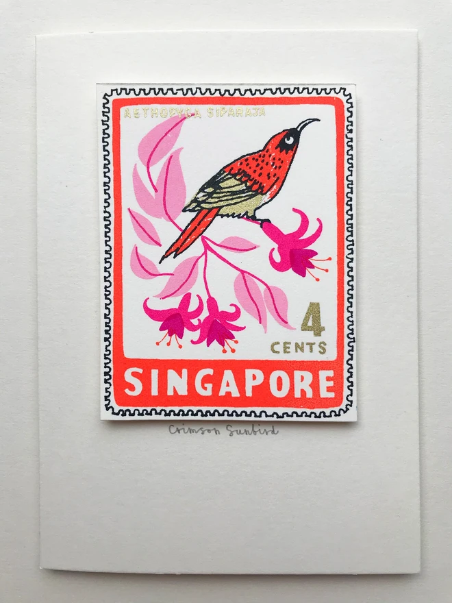Bird Stamp Card Singapore/ Crimson Sunbird, a white card with a colourful bird design in the centre on top of a white surface. 