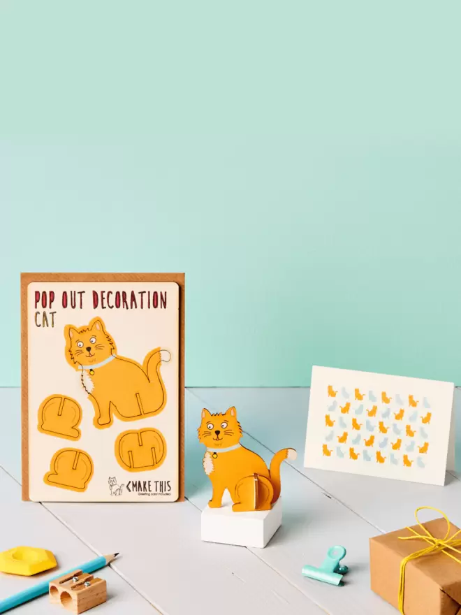 3D ginger cat decoration and cat pattern greeting card and brown kraft envelope on top of a grey desk in front of a light green coloured background