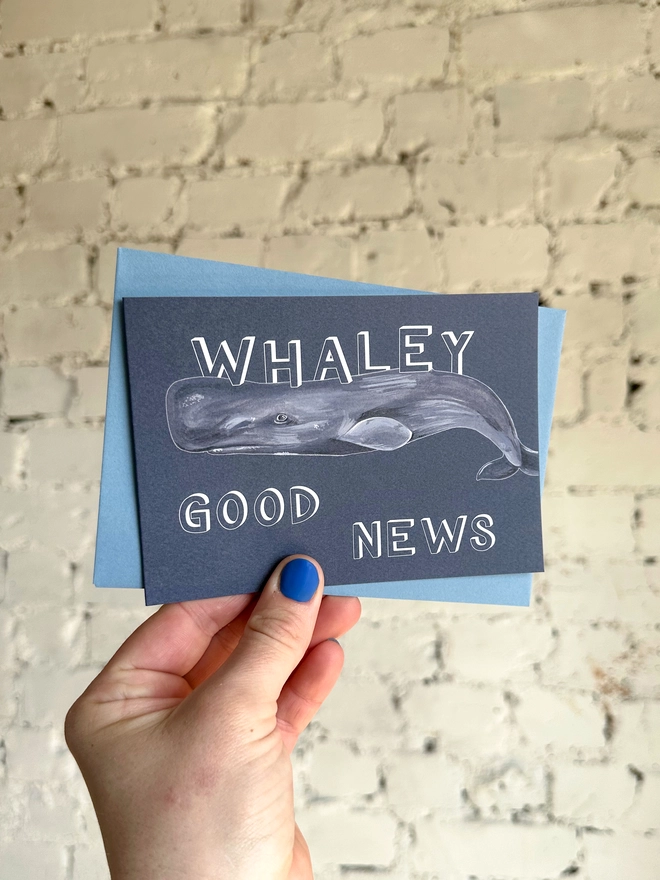 a dark grey blue greetings card featuring an illustration of a sperm whale with the text “whaley good news” with a blue envelope