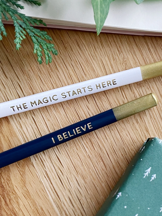 Two pencils, one white and one navy blue, both with gold writing along the sides, lay on a wooden desk.