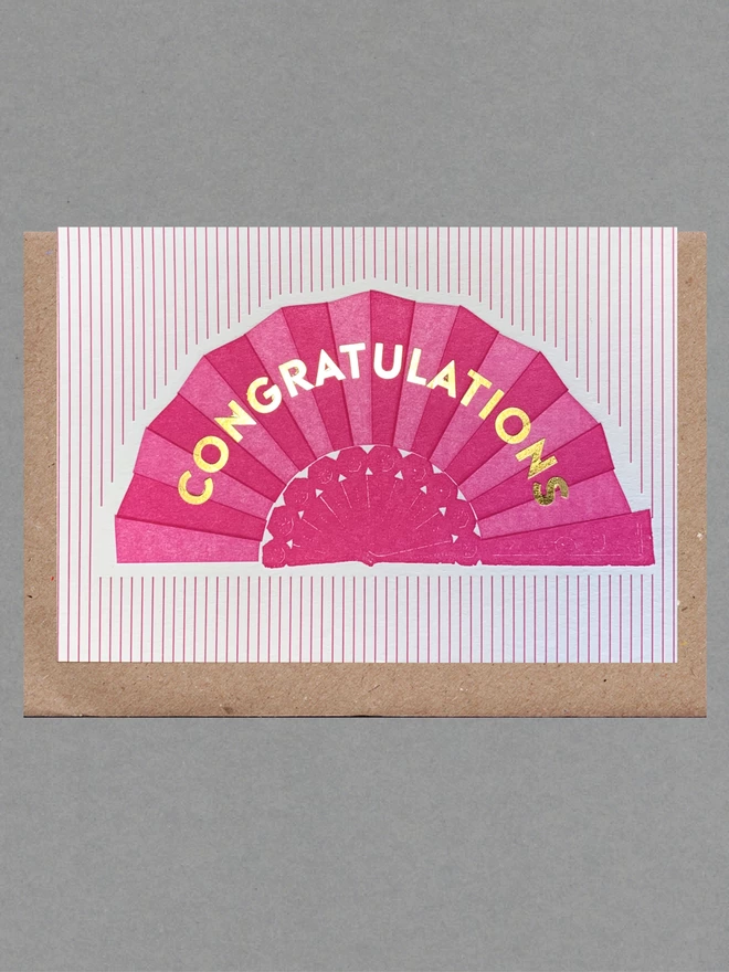 Pink and white striped card with a pink fan on it and gold text reading 'Congratulations' with a brown envelope behind