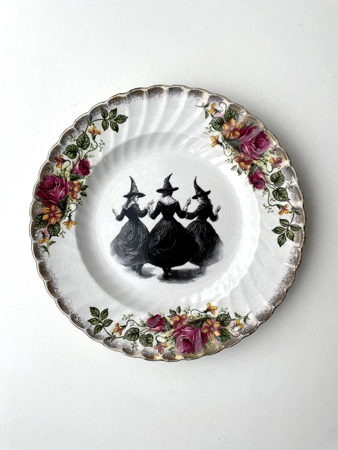 a coven of 3 witches dance hand in hand, the image is victorian in black and white and is printed directly onto an ornate vintage plate