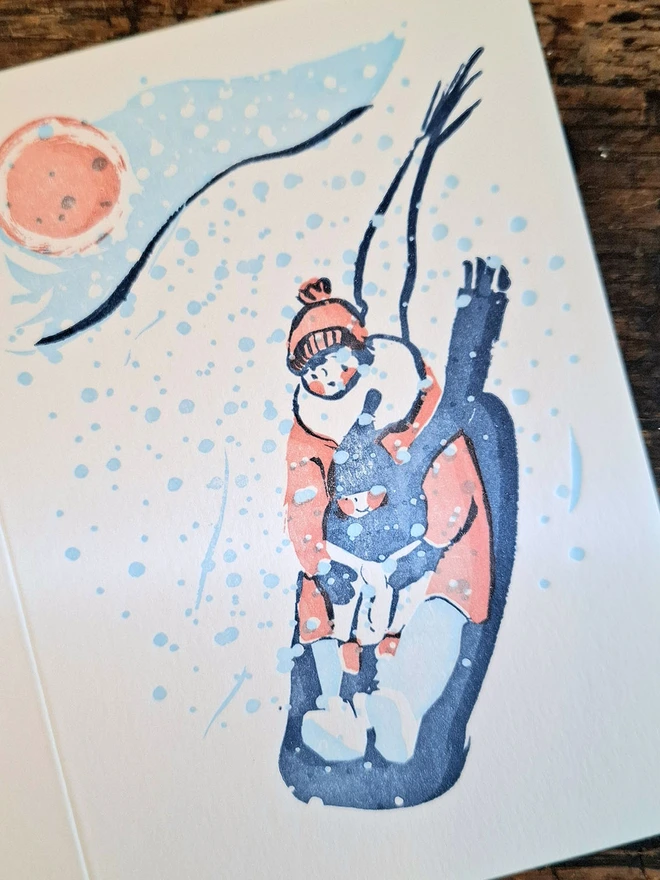A handprinted greetings card lying open with the image facing up on a wooden surface.  The illustration on the front of the card is printed in pink, light blue and dark blue and is of a mother holding her young child as they sledge down hill in snow.