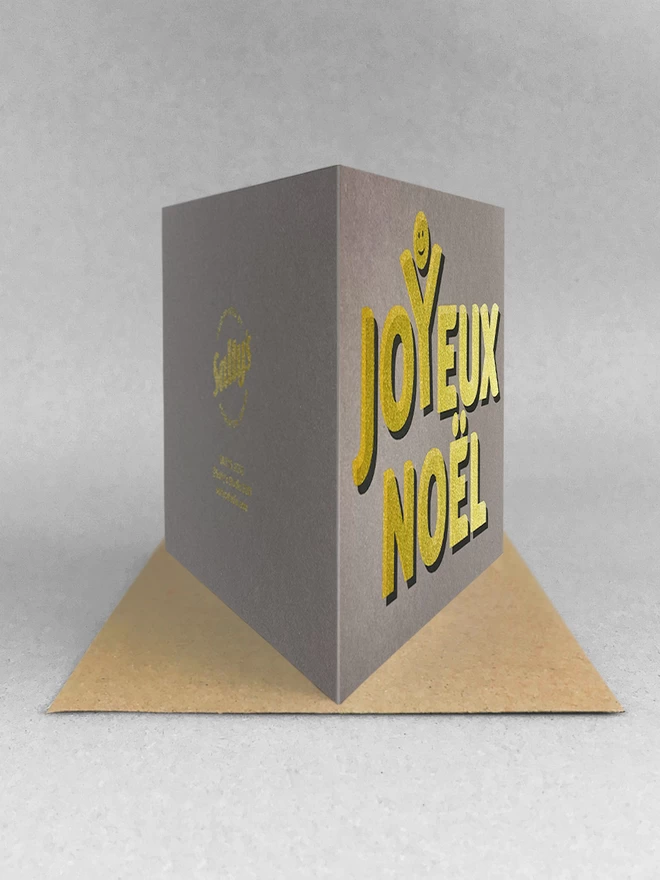 Golden text screenprinted onto grey card stock, spells out Joyeux Noel with a happy face above the Y, as though celebrating. Landscape card stood on a grey background, stood on a kraft brown envelope viewed from the rear.