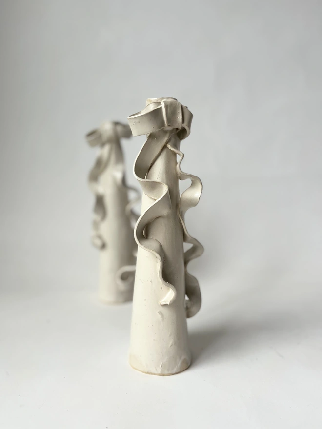 two cream white stoneware tall candlestick holders with a ceramic bow with long ribbons flowing down the body of the candlestick itself