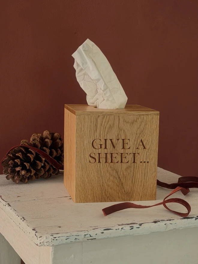 Oak tissue box cover engraved GIVE A SHEET..., on a white painted stool against a red wall