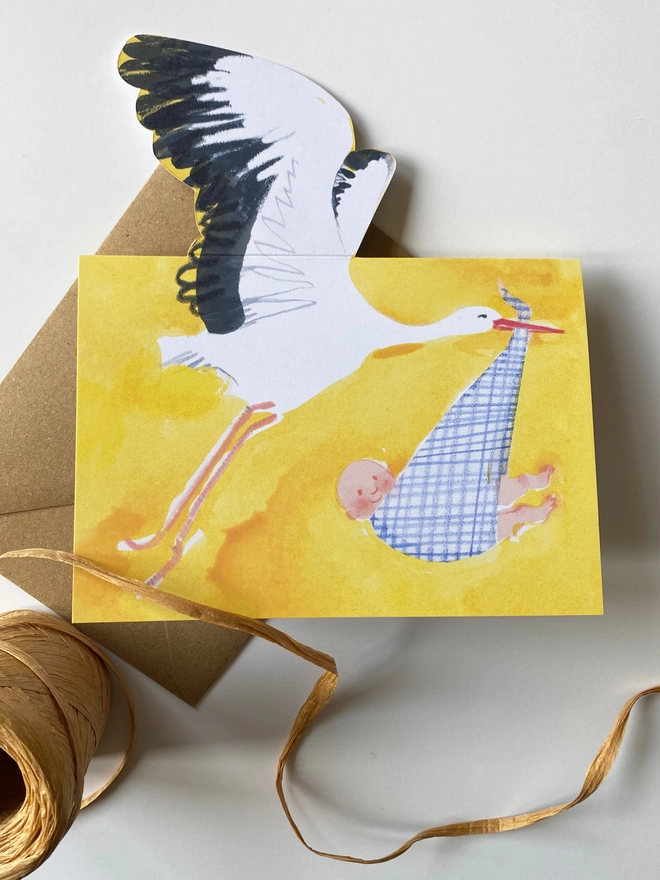 A new baby card by Esther Kent showing a black and white stork carrying a baby in a blue checked cloth, on a yellow background