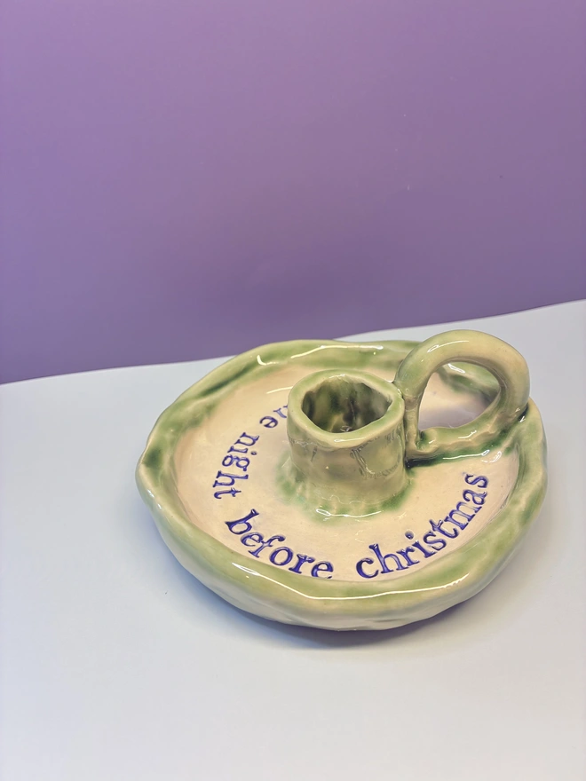 Handmade ceramic Christmas taper candlestick holder with a soft green glazed edge and the words “‘Twas the night before Christmas” in dark blue lettering, arranged around the candle holder opening. Photographed against a pale background.