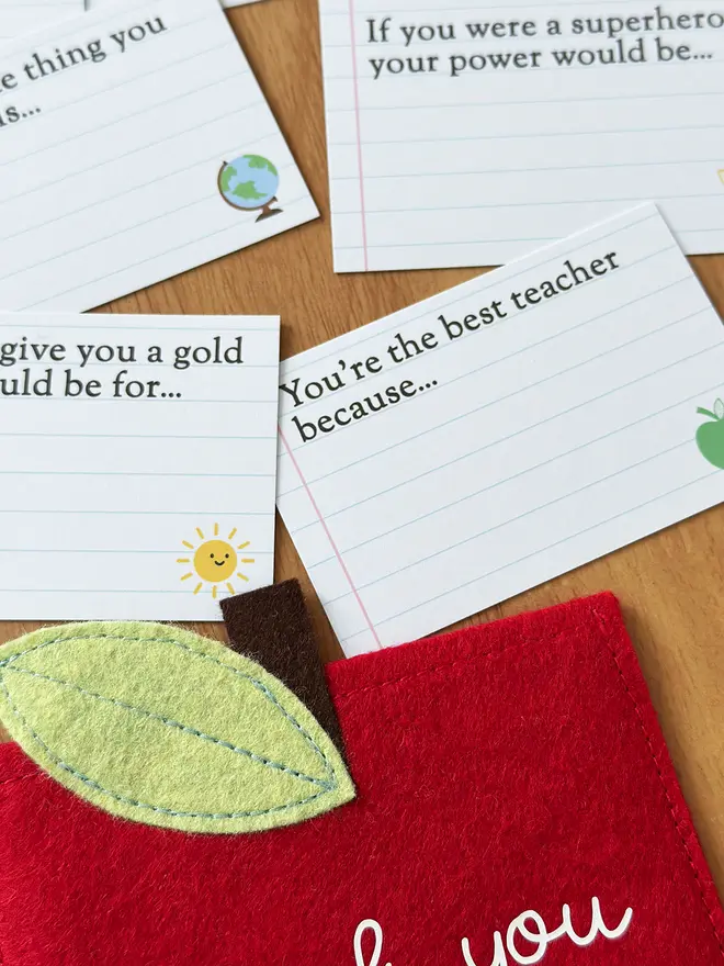 A handmade red felt apple-shaped envelope with "Thank you Miss March" written on the front. The heirloom apple envelope surrounded by completed prompt cards with handwritten messages from a child, displayed on a wooden desk. It's a thoughtful personalised gift for teachers from children.