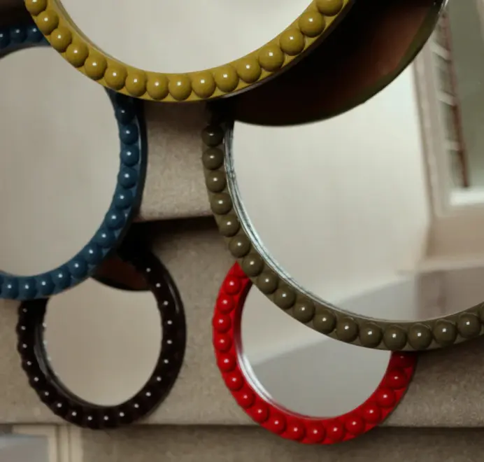 Many round oversized bobbin mirrors, in different colours and sizes with a decorative bobbin-style wooden frame, hanging on a white wall.