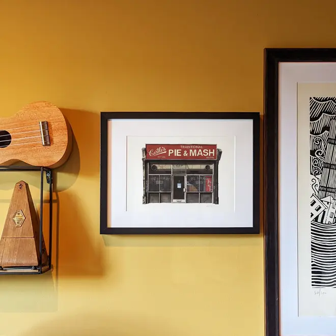 Camden Castle's Pie & Mash Shop Giclée Print (Unframed), a print of a shop sitting against a mustard backdrop. It is accompanied by other framed prints. 