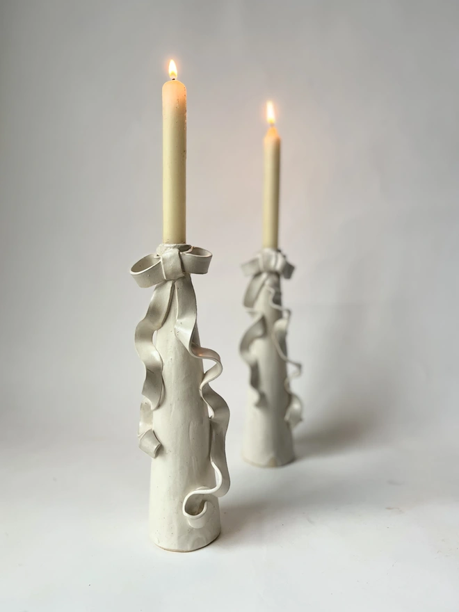 two cream white stoneware tall candlestick holders with a ceramic bow with long ribbons flowing down the body of the candlestick itself, and two lit candles in the holders