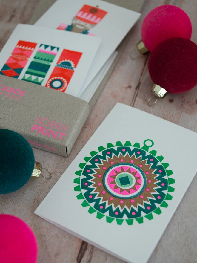 Three mini christmas cards tumble from their box, this set of mini Christmas cards is surrounded by small brightly coloured baubles.