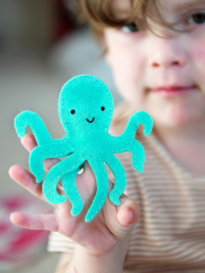 A young child is holding a turquoise felt octopus finger puppet on their fingers.