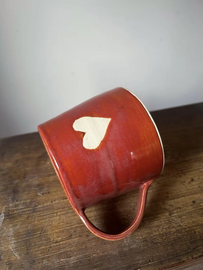 Heart Mug Cinnamon, a heart mug on a wooden surface lying on its side in front of a plain background. 