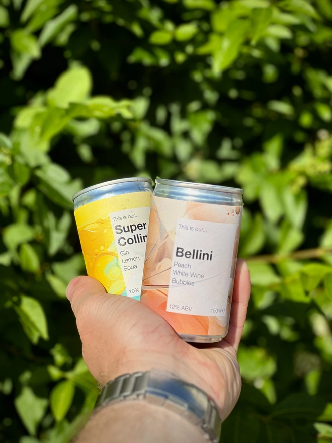 Bellini and Super Collins cans in a hand, photographed against a green bush