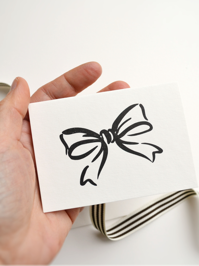 Small greeting card with simple bow design in the palm of a hand.  Letterpress printed with black ink on a thick cream paper.