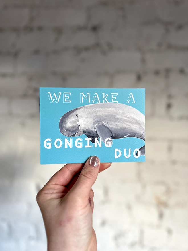 a bright blue greetings card featuring an illustration of a dugong with the text “we make a gonging duo” 