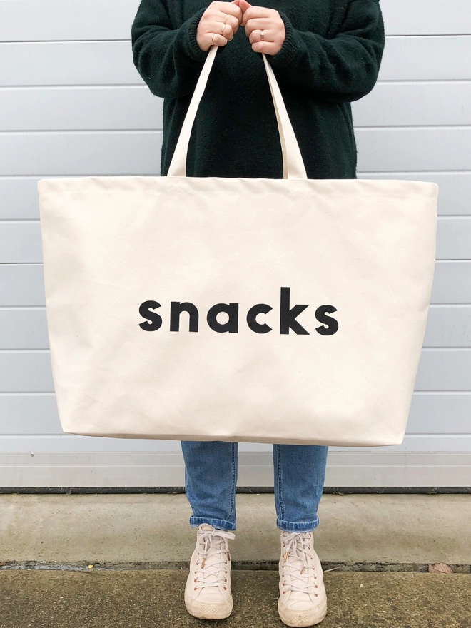 model holding an oversized natural canvas tote bag with bold snacks slogan