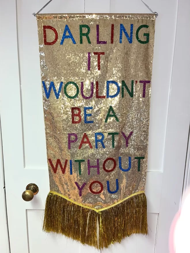 A midi customisable banner hangs from a hook. It has a gold sequin background and a gold tinsel trim along the bottom. The text is multicoloured and says 'DARLING IT WOULDN'T BE A PARTY WITHOUT YOU'