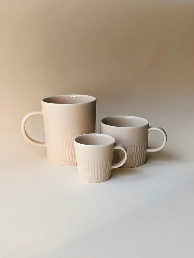 Three handmade porcelain mugs and coffee cups in a Neutral Pale Oat. A large tea mug with a hand carved scalloped texture around the base, a small mug for flat white coffee which is hand carved in a combed texture, and espresso cup with scalloped hand carving around the base. All are glazed inside and matte outside.