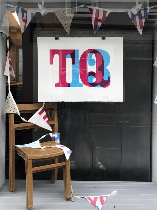 Our shop window with a village hall chair stood on a grey wooden floor, bunting trailing from above to the floor and a print clipped in position. It is a print of big postcode letters and numbers screenprinted in magenta and cyan ink overlapping somewhat.