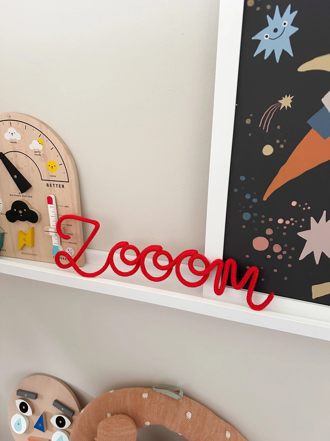 A kids wall sign displayed on a shelf with the word 'zooom' for an astronaut or space themed room. Shown next to a space themed print and other wooden childrens toys. 