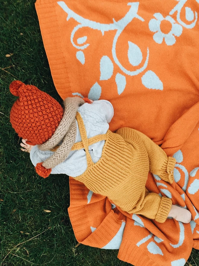 A close up of a baby in warm knitted dungarees and bonnet in autumn colours crawling on the rust briar rose baby blanket.