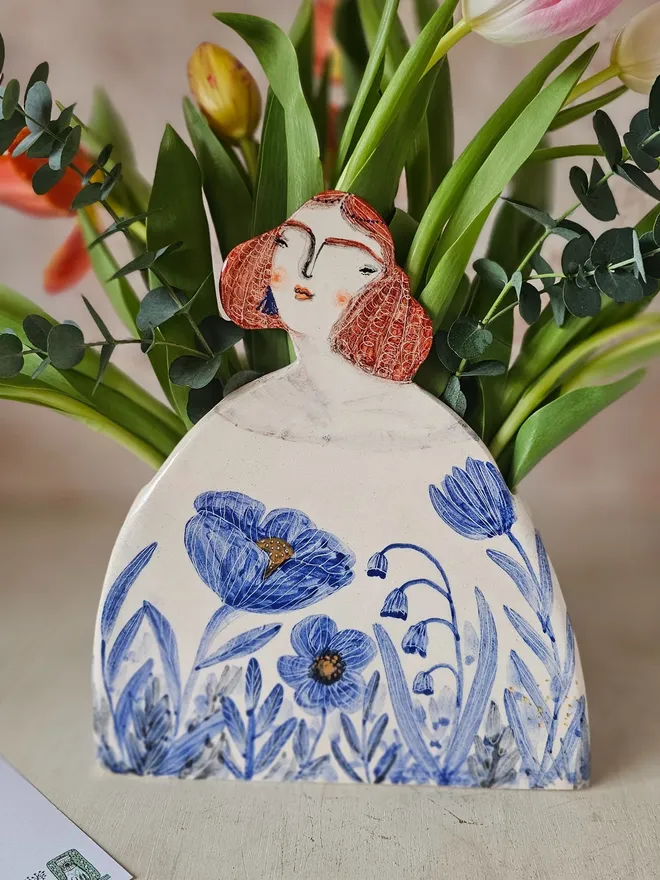a large vase shaped like a lady with red hair looking up with blue flowers painted on her dress, front view with flowers