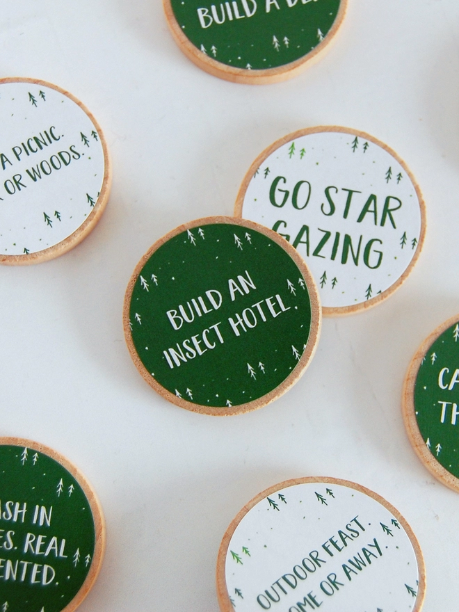 Several wooden tokens with white and green labels lay on a white wooden surface. Each token has an outdoor adventure idea printed on.