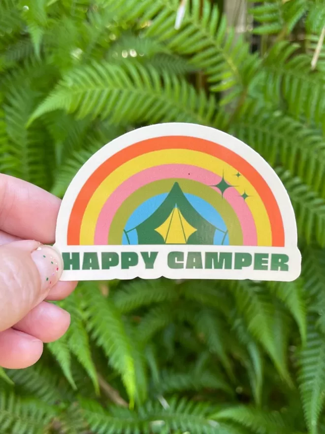 A happy camper rainbow vinyl sticker being held up in front of plants.