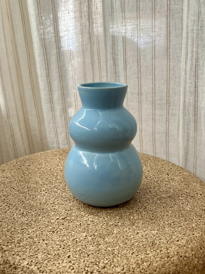 sky blue chubby bud vase sitting on a cork table with a curtain in the background. The bud vase has curvy sides with a flared rim 