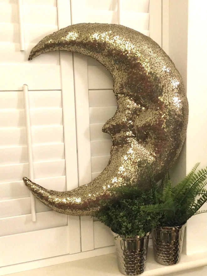 A large gold sequin plush half crescent moon sits on a white shuttered window sill  above pot plants.