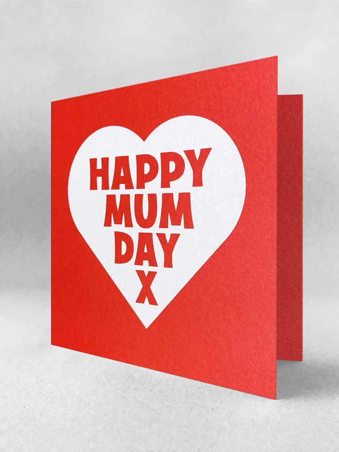 A red square card with a white heart, containing the words Mappy Mum Day x. Stood slightly open in a light grey studio 