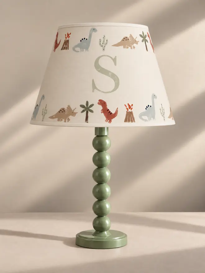 Personalised dinosaur lampshade with initial S, hand-painted with colourful dinosaurs, palm trees, cacti and volcanoes on a green table lamp.