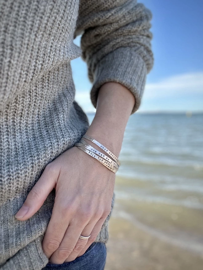 A woman in a green knitted jumper and blue jeans, wearing a stack of silver cuff bangles, standing in front of the sea with blue skies in then background.