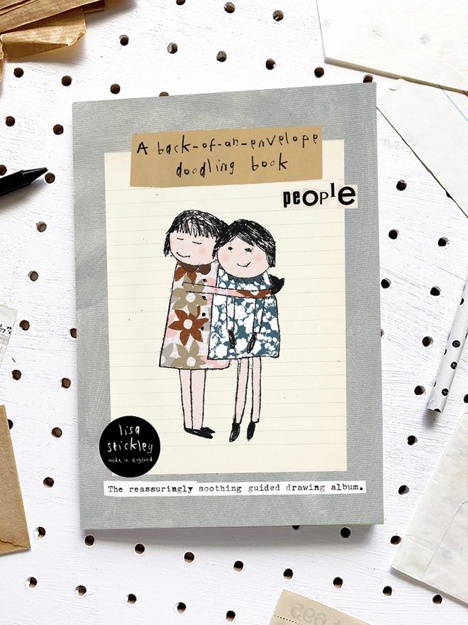 sketchbook on white pegboard with pale grey cover and illustration of two girls hugging