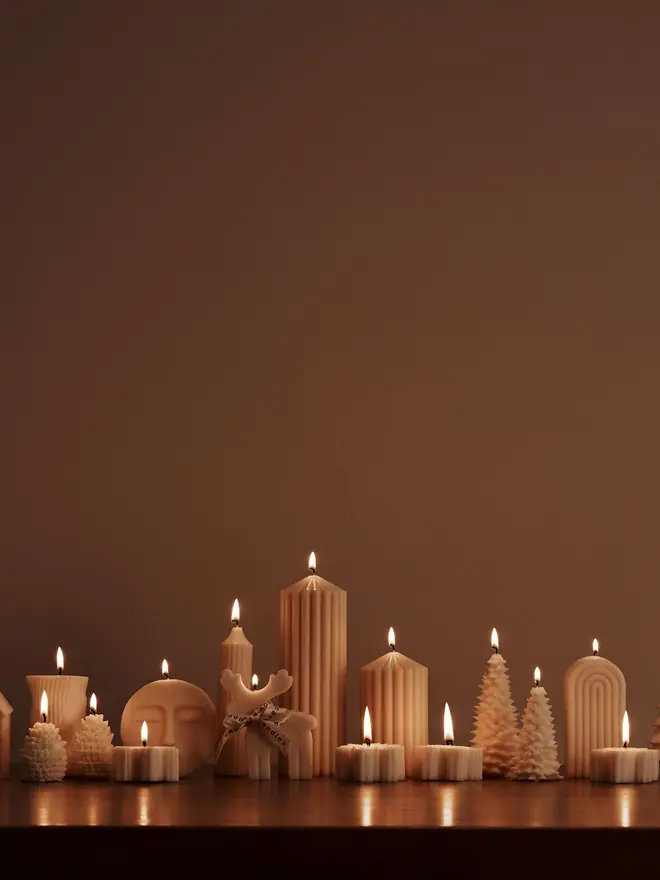 Reindeer Sculpture Candle. A row of lit cream sculptural candles, including a reindeer, house, Christmas trees, a moon face, an arch and ribbed pillars, on a dark wooden mantelpiece. The warm glow of the collection creates a cosy, atmospheric festive display