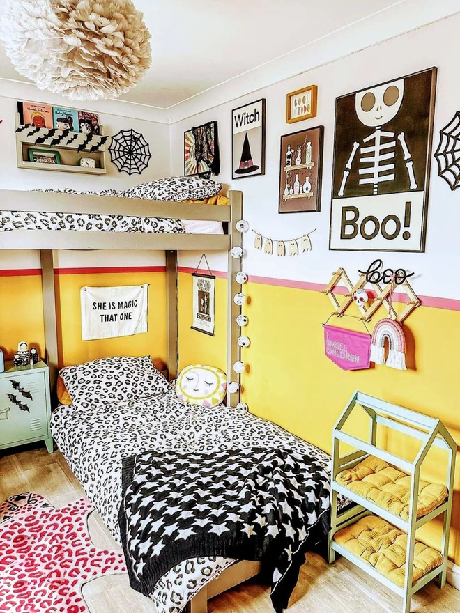 A modern shared bedroom dressed for Halloween, with  a few cute spooky posters of skeletons and spiders webs, and a black and white star junior blanket draped across the bottom bun  bed.