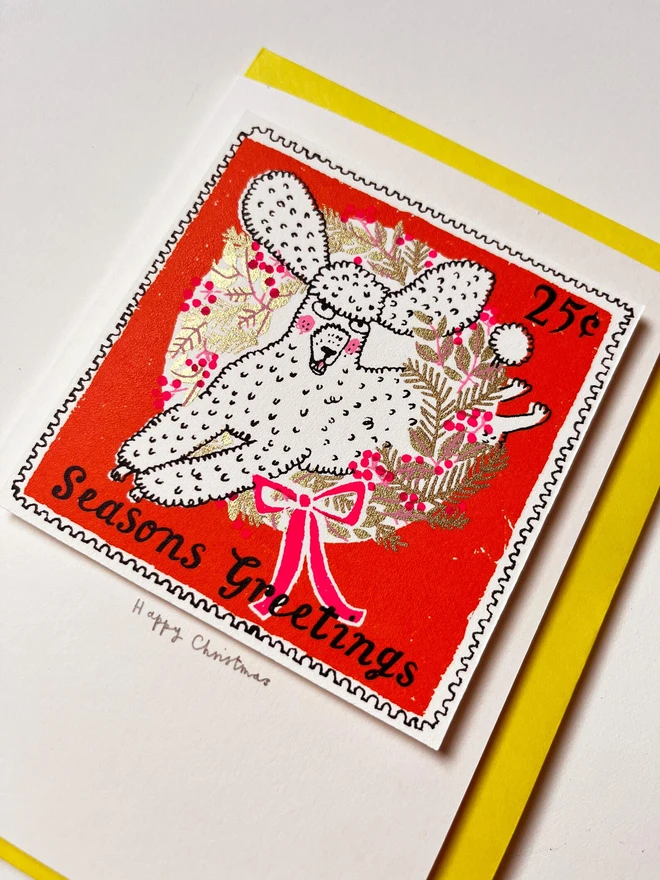 Christmas Poodle Card, a Christmas card with a colourful design in the centre on a yellow envelope. They are placed on a white surface. 