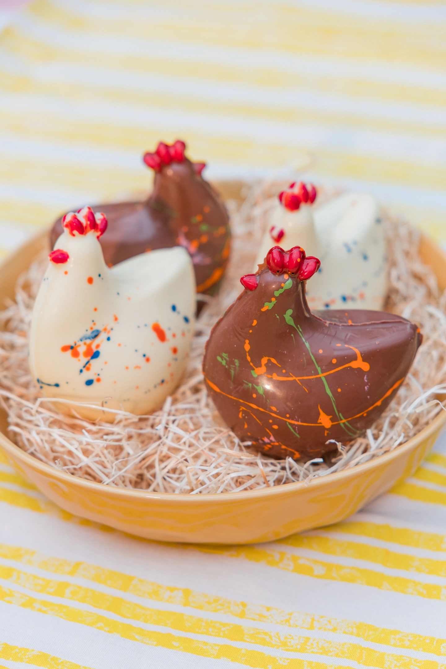 A yellow bowl filled with straw, sat 4 chocolate chickens. Two white chocolate, two milk chocolate. On a table with a striped yellow and white tablecloth. 