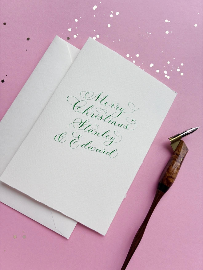 Christmas card in handwritten green calligraphy with a wooden penholder and pink glitter background