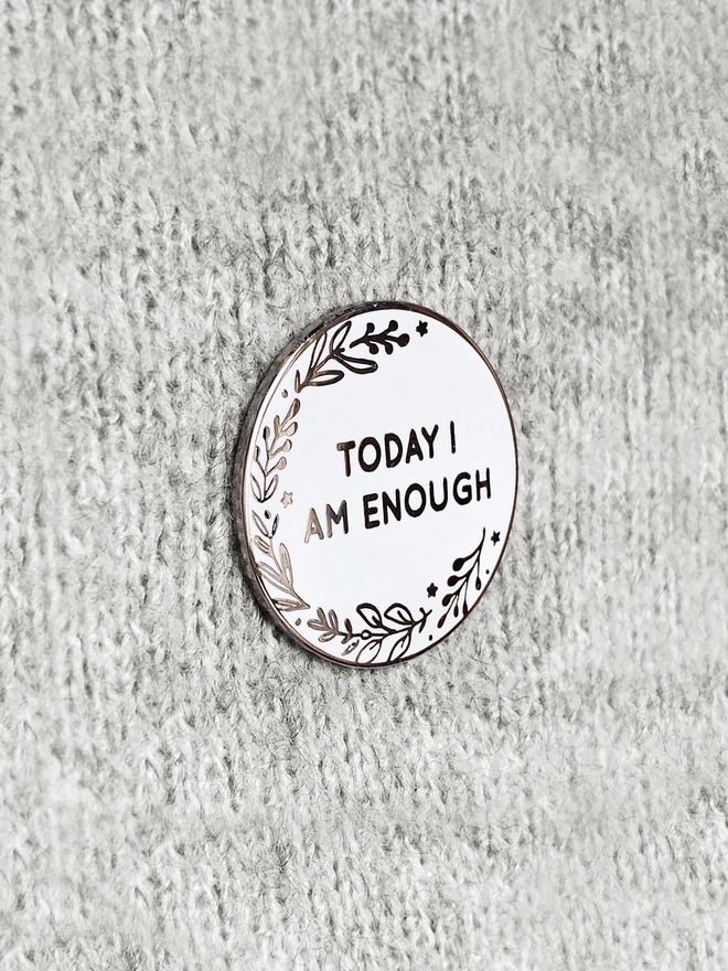 A round white enamel pin with a botanical design and the words "Today I Am Enough" is pinned to grey fabric.
