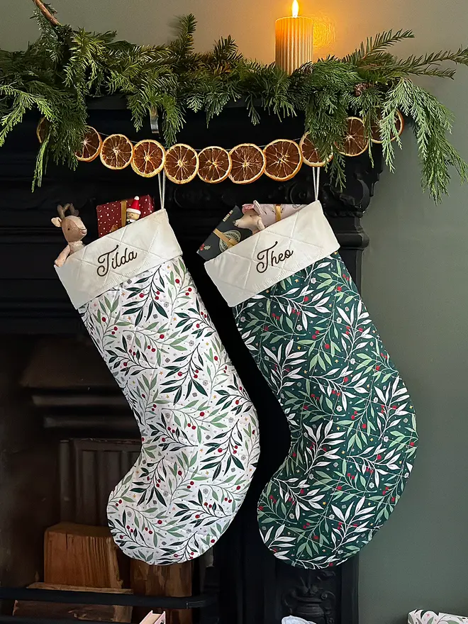 Two handmade quilted Christmas stockings hang on a fireplace decorated with greenery and orange slice garland.